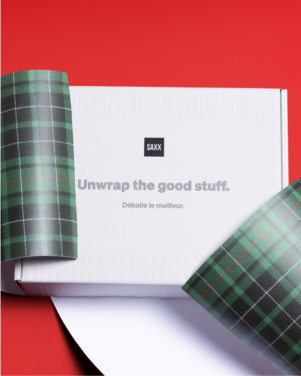 A white square box with the SAXX logo and the text 'Unwrap the good stuff.' on top, photographed on a bright red background, with green wrapping paper.