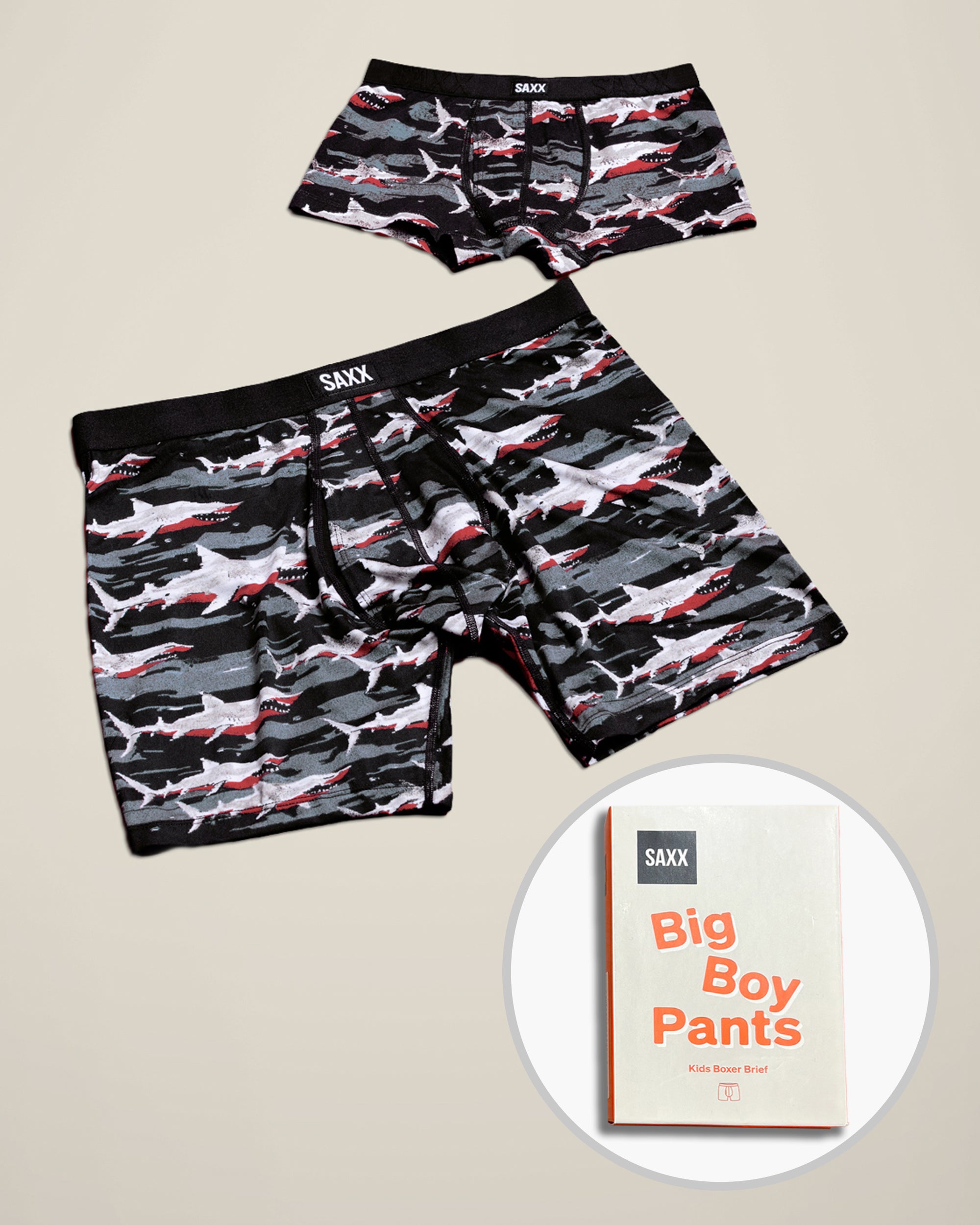 Two pairs of SAXX Father/Son boxer briefs with a shark camouflage pattern in black, gray, and red, displayed on a beige background with a circular inset showing a box with 'Big Boy Pants'