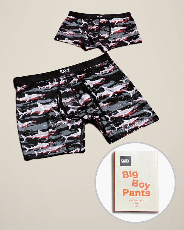 Two pairs of SAXX Father/Son boxer briefs with a shark camouflage pattern in black, gray, and red, displayed on a beige background with a circular inset showing a box with 'Big Boy Pants'
