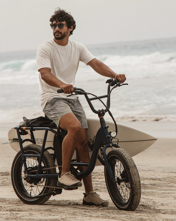 Man wearing patterned swim shorts while riding a electric bike on a beach with a surfboard attached 
