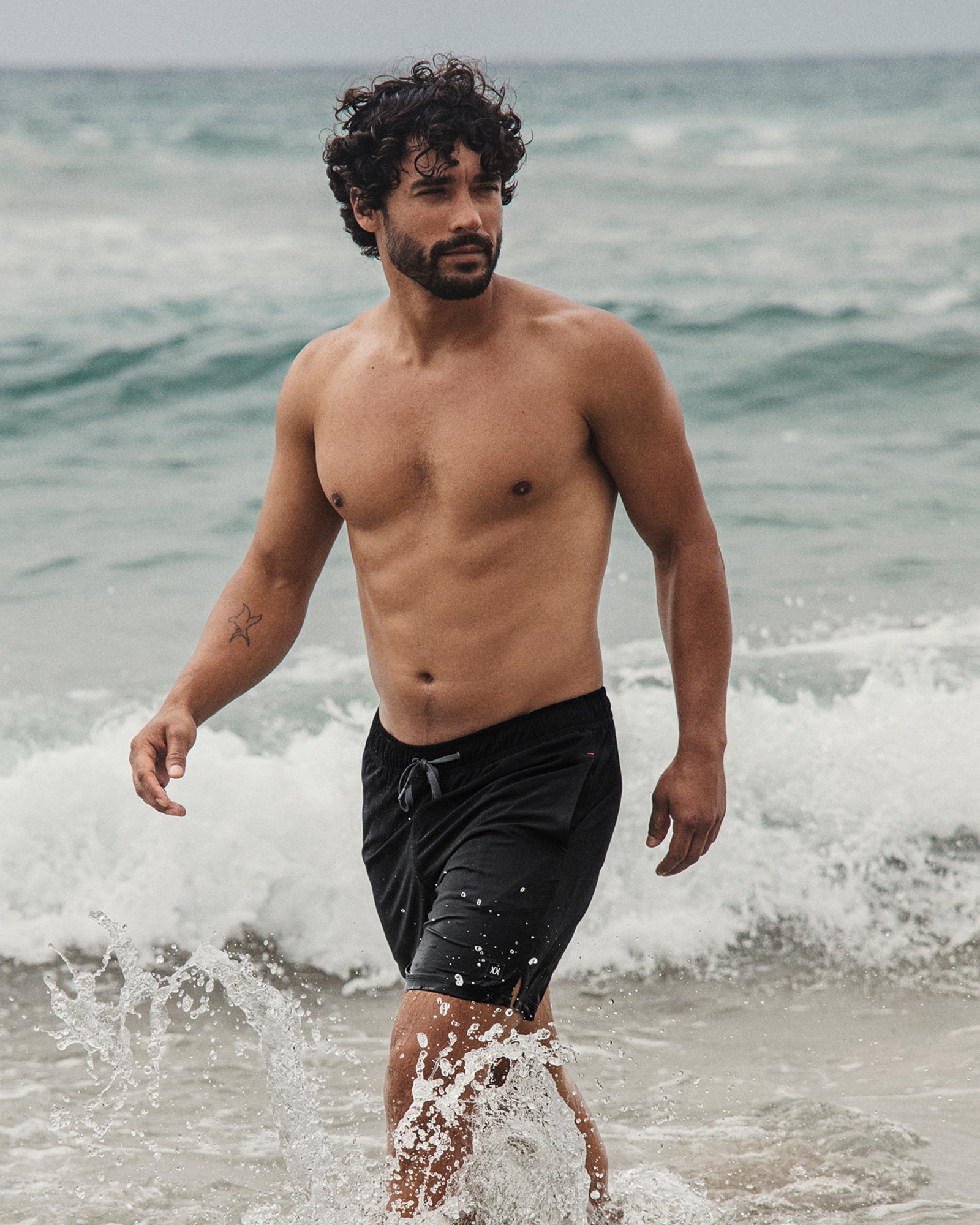 Man walking in the ocean wearing black swim shorts with waves around him