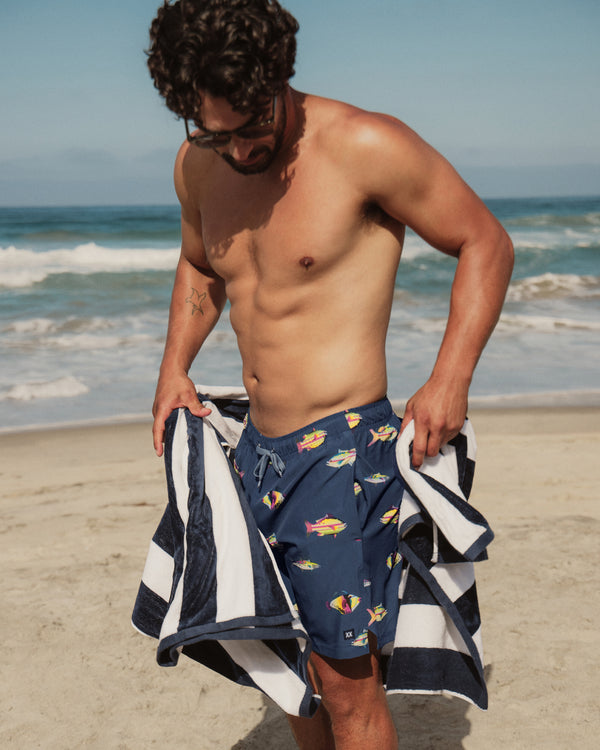 Man on a beach wearing navy swim trunks with colorful fish pattern, holding a striped towel.