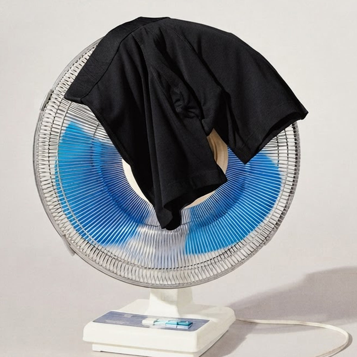 A black short-sleeve T-shirt draped over a small white pedestal fan with blue fan blades, photographed against a plain light background.