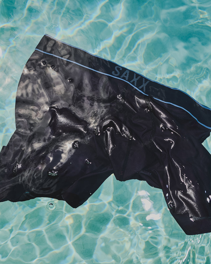 Black wetsuit or swim trunks with a glossy finish and a light blue waistband stripe, floating on clear turquoise pool water. The brand name 'SAXY' is visible on the waistband.