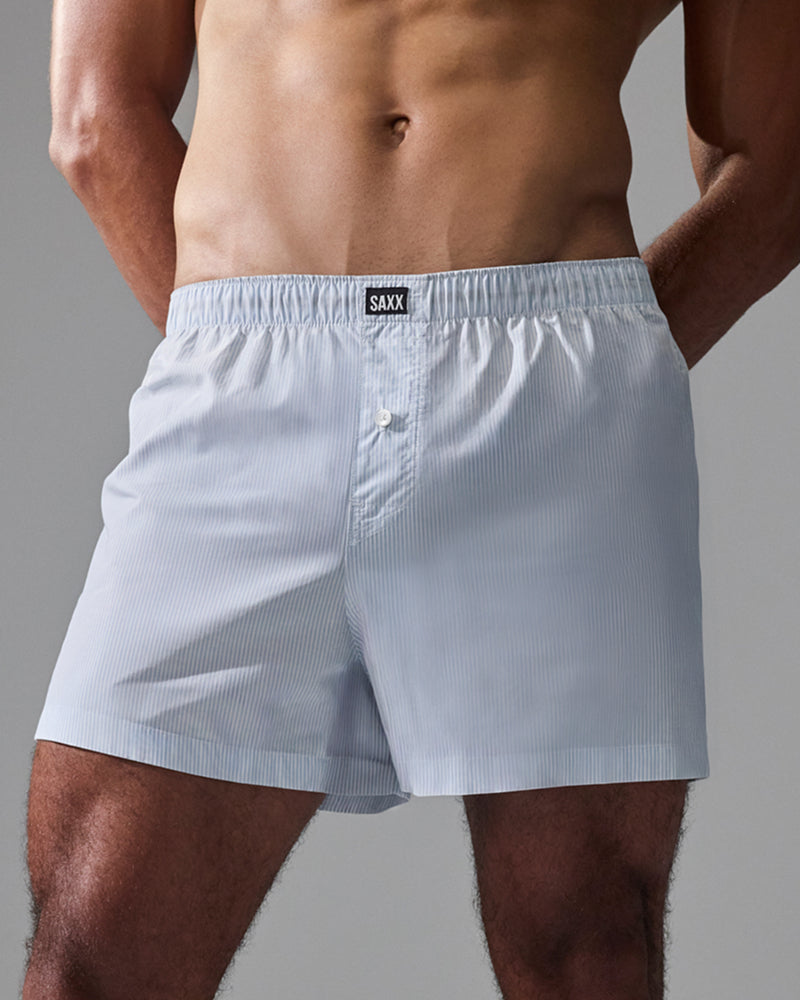 A close-up of a man wearing light blue pinstriped boxer briefs with a visible SAXX waistband label, shown against a plain gray studio background.