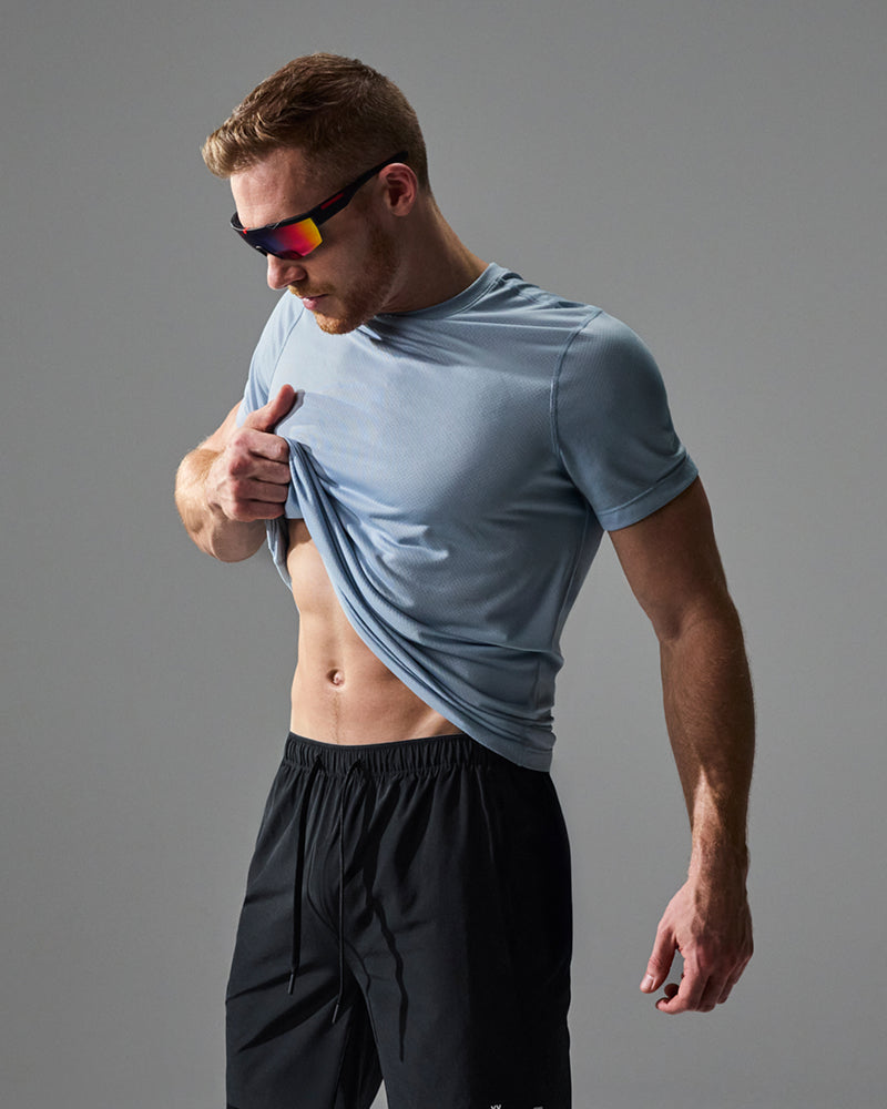A man wearing a light blue short-sleeve athletic t-shirt and black shorts, lifting the shirt to show its fit. He is wearing black sunglasses with colorful mirrored lenses against a solid gray studio background.
