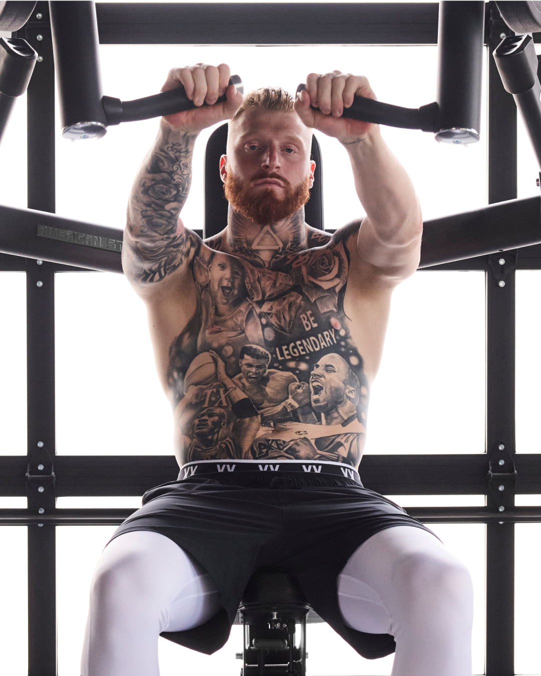 Pro football player Maxx Crosby sits on a black gym machine, gripping the handles above his head. He is wearing black athletic shorts and white compression sleeves.