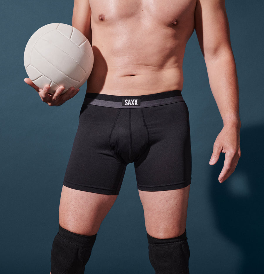 Man holding volleyball while wearing knee pads and black boxer briefs