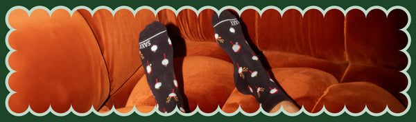 Man wearing black socks in a reindeer print with his feet on a red velvet couch