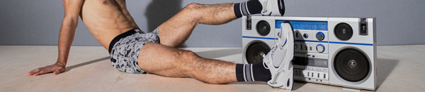 Shirtless man in grey underwear and shoes with feet up on boombox