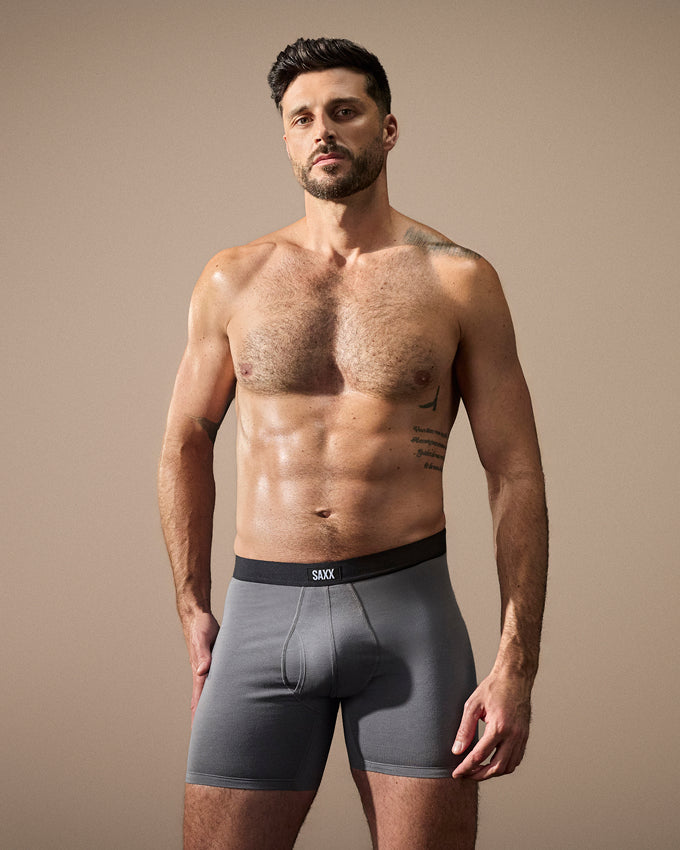 A shirtless male model wearing gray boxer briefs with a black waistband featuring the brand name 'SEXX', posed against a solid beige studio background.