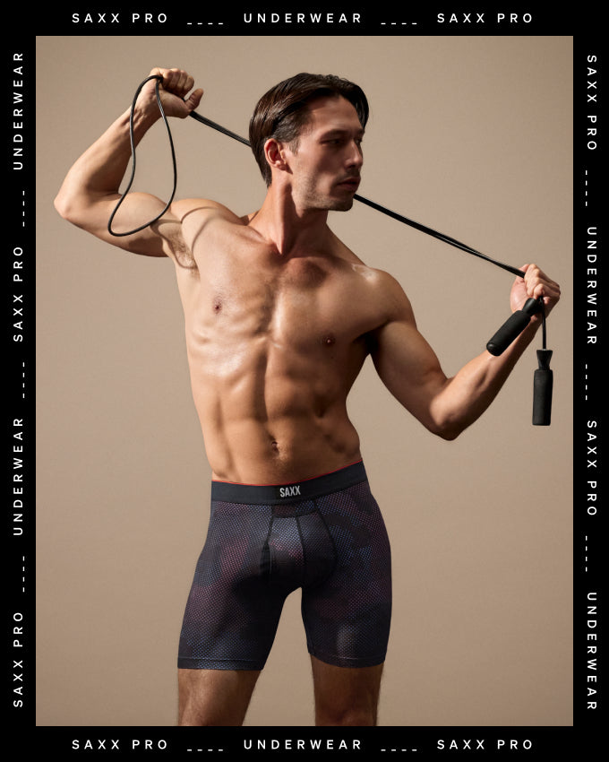 A muscular man modeling SAXX PRO underwear, holding a jump rope against a warm beige studio background. The brand name 'SAXX PRO' is repeated along the borders of the image.