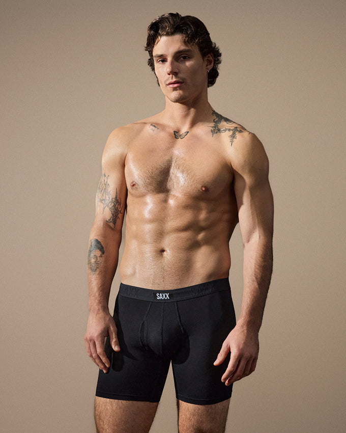 A shirtless male model wearing black SAXX boxer briefs, standing against a solid beige studio background.