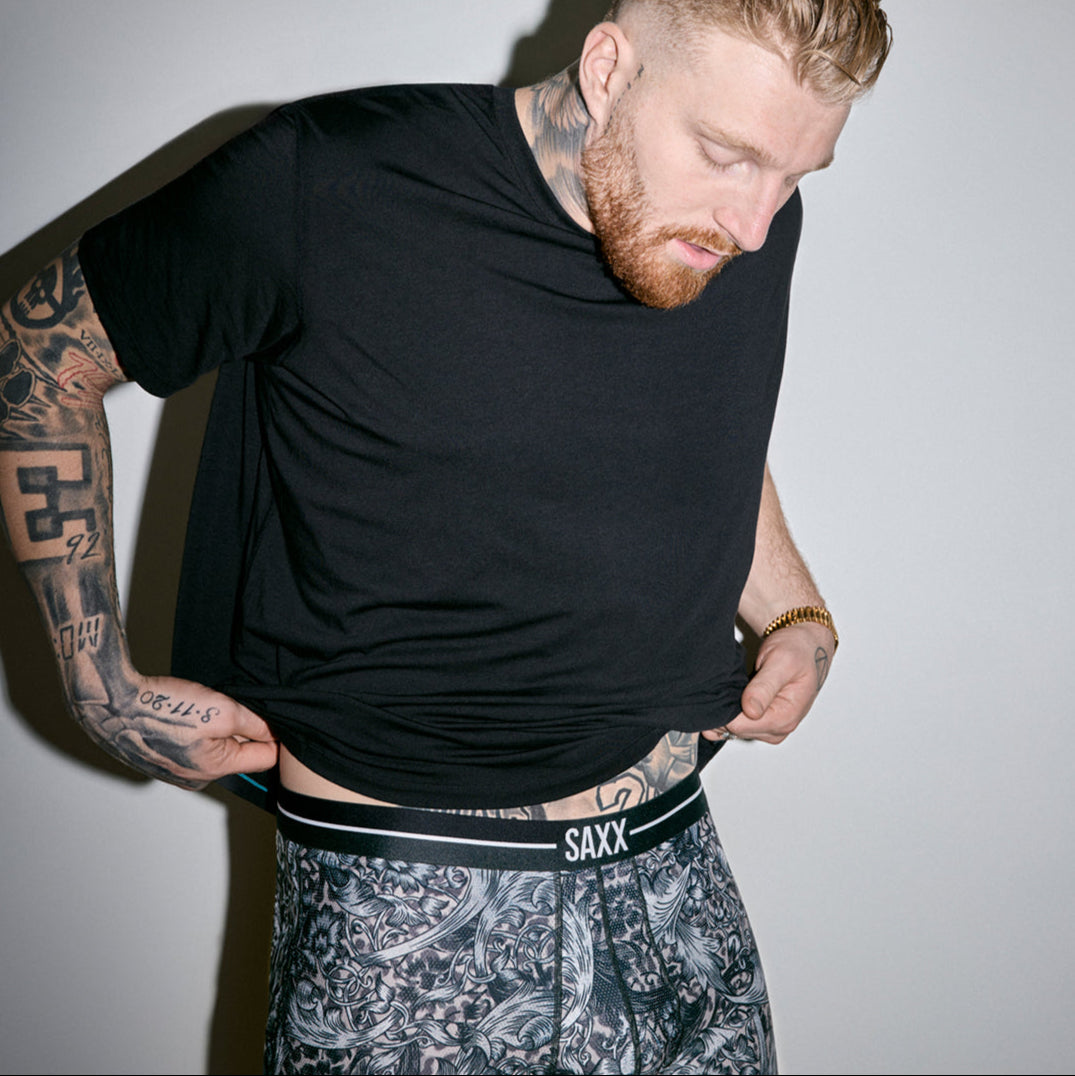 A heavily tattooed man lifts up his shirt to expose the waistband of his boxer briefs.