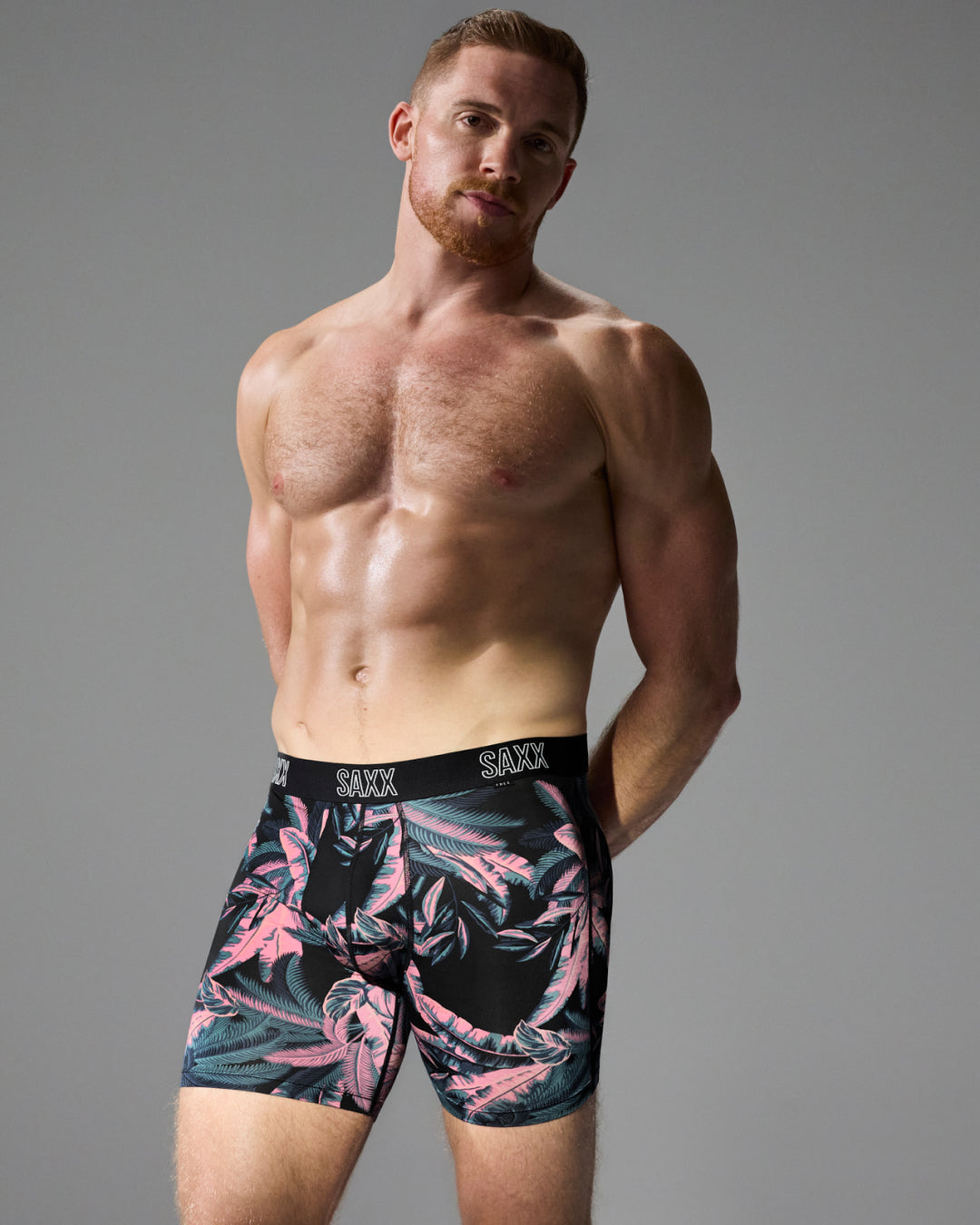 A shirtless male model wearing SAXX tropical print boxer briefs with a black waistband, photographed against a solid gray studio background.