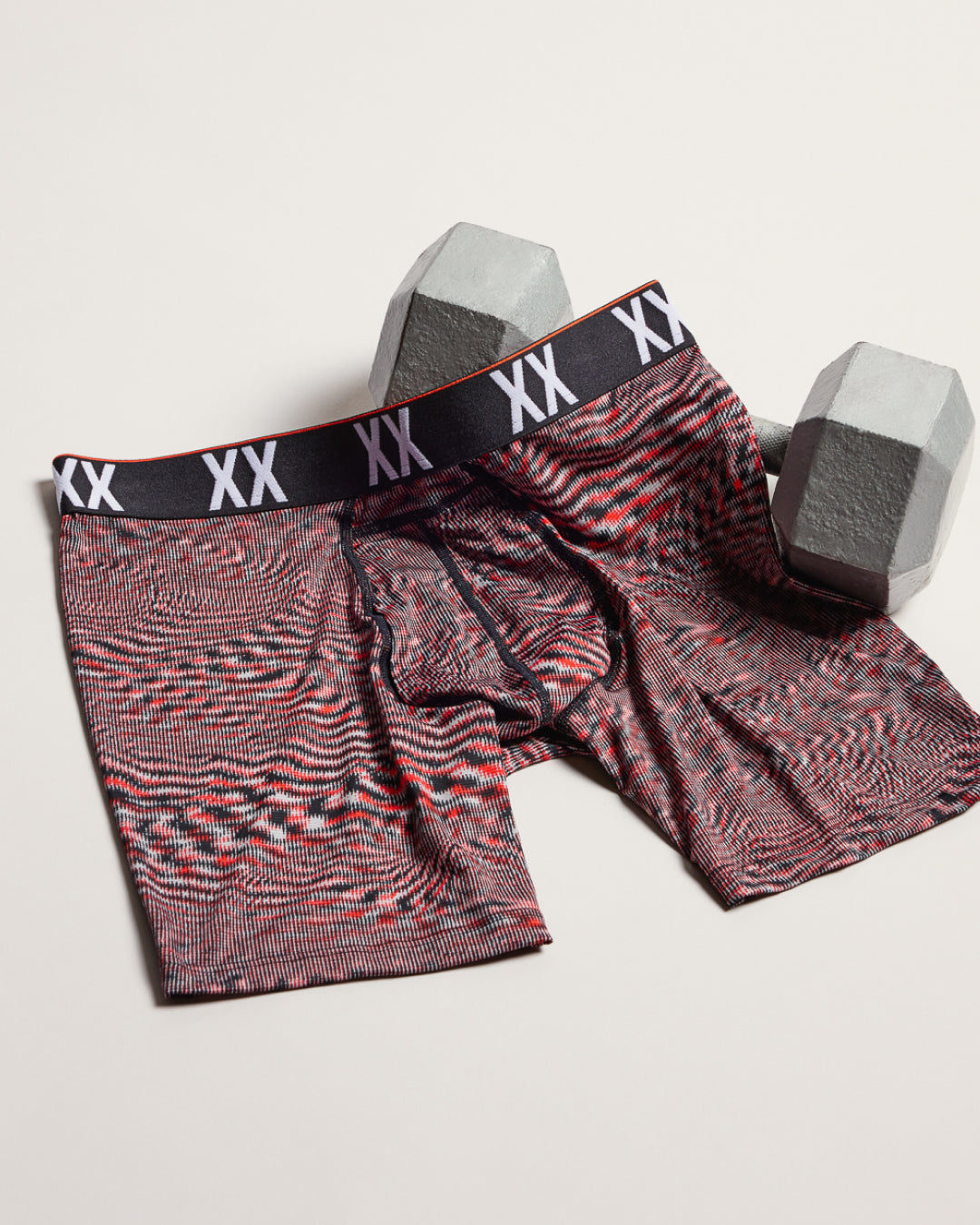 A pair of XX brand boxer briefs with a red, black, and gray wavy pattern, displayed on a light beige background with two gray hexagonal dumbbells behind them.