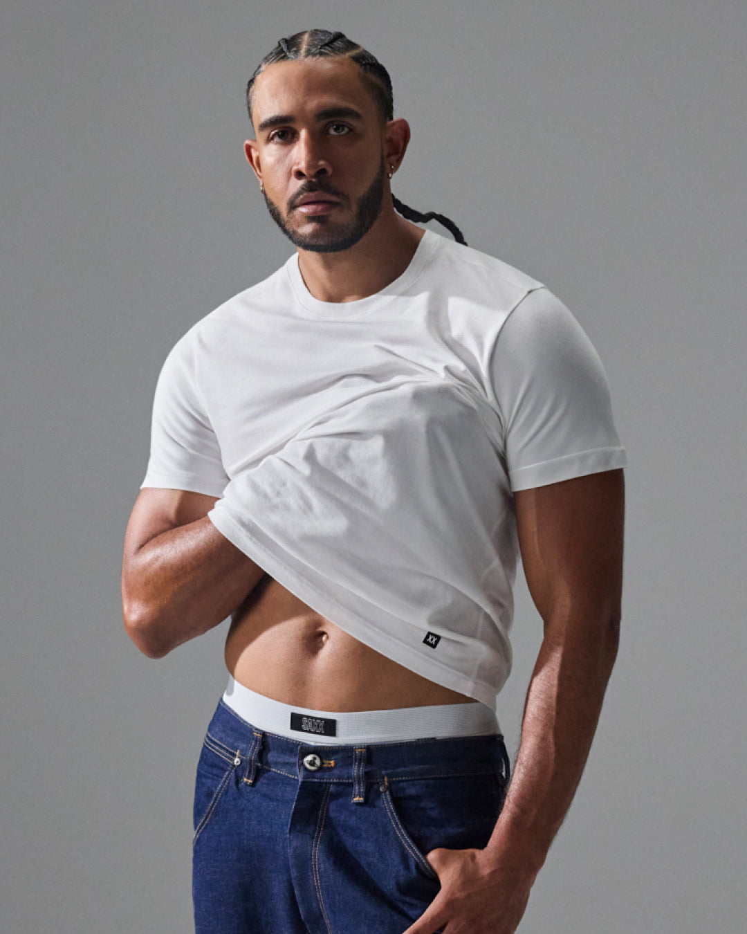 A muscular man wearing a white short-sleeve crop top t-shirt and blue jeans, posing against a solid gray studio background.