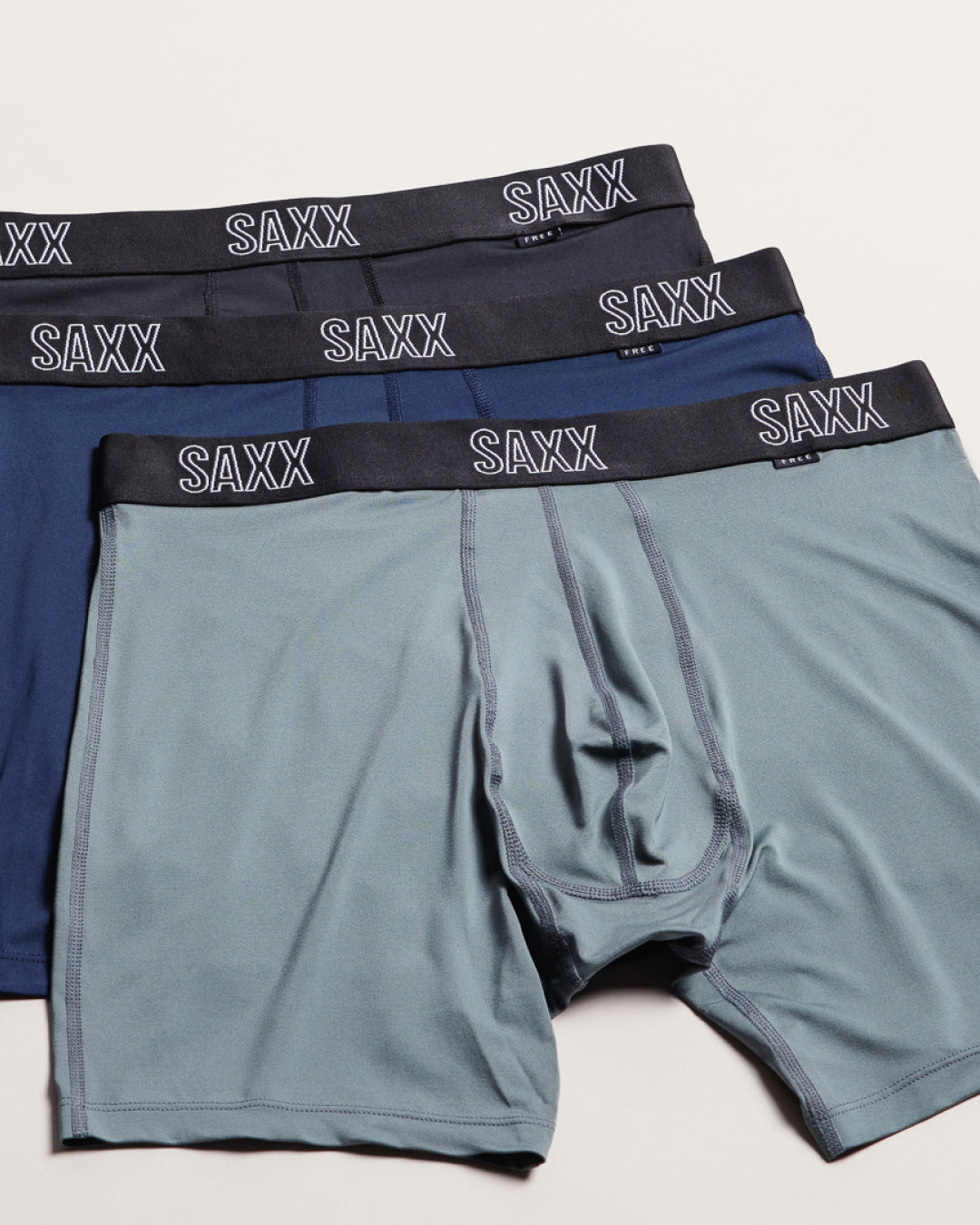 Three pairs of SAXX men's boxer briefs stacked diagonally, featuring a slate blue front pair, a navy blue middle pair, and a dark charcoal gray third pair, all with white brand lettering on the elastic waistband.