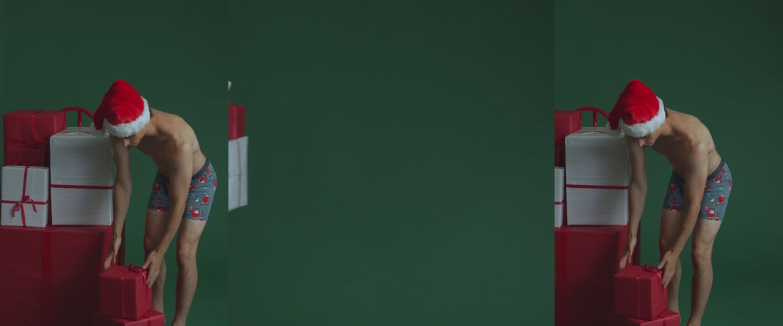 Shirtless man in festive SAXX Underwear print and Santa hat leaning over and grabbing gift from pile of presents beside him