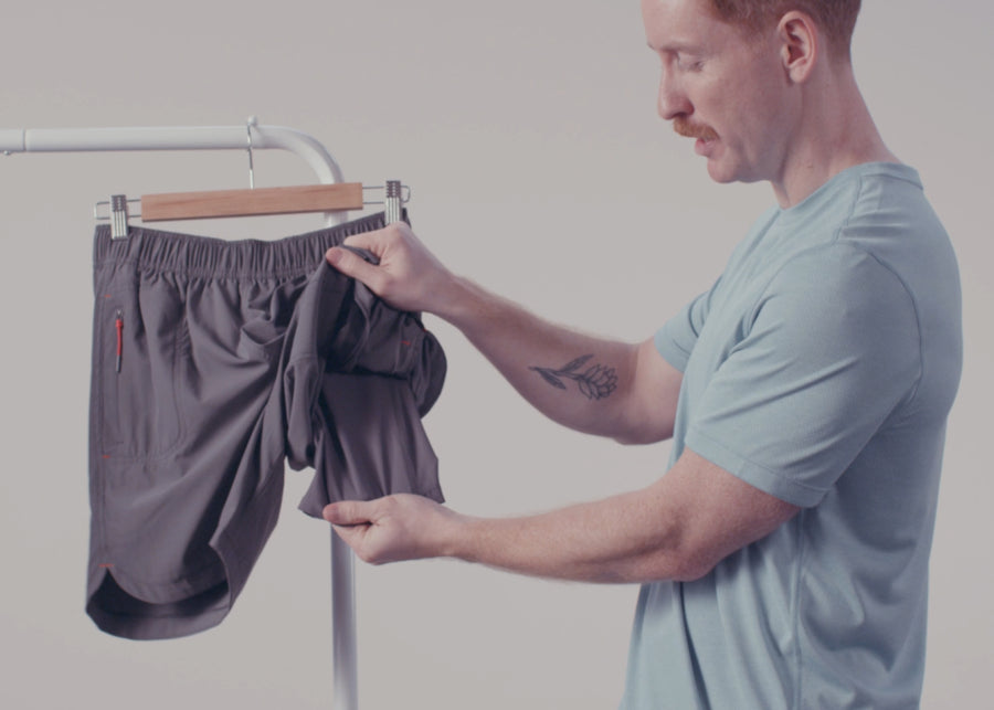 A man in a grey SAXX tee showing the liner in a pair of grey SAXX Gainmaker Shorts hanging on a rack. 