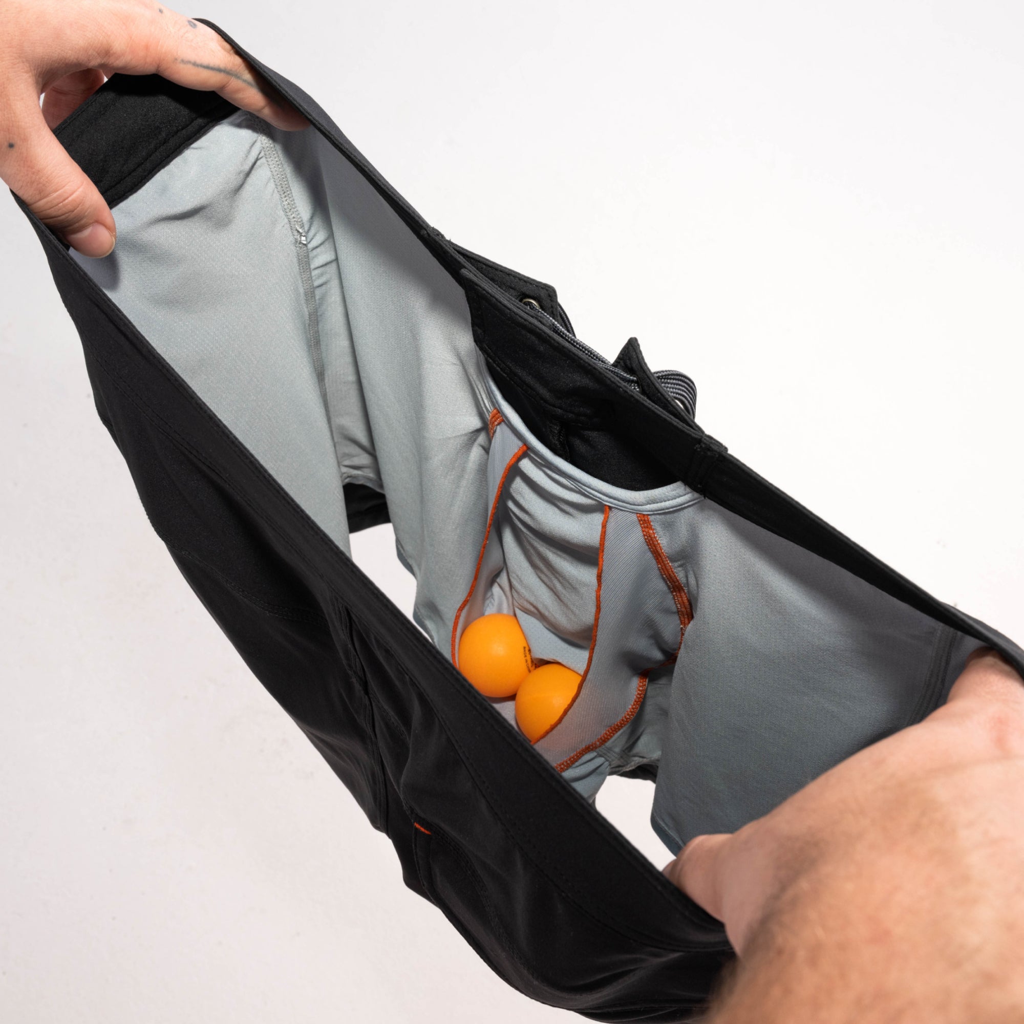 Man's hands revealing inside of 2N1 shorts showing liner with pouch holding two ping pong balls
